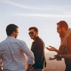 Tres amigos riendo y conversando al atardecer. Uno dice: "Encantado de conocerte en inglés", mientras los otros dos escuchan atentamente.