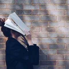 Persona con un libro abierto cubriendo su rostro, apoyada contra una pared de ladrillos. Parece estar inmersa en la lectura, quizás buscando un momento de tranquilidad.