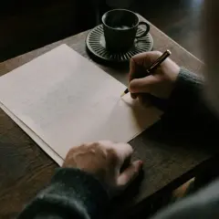 Persona escribe una carta en inglés, con una taza de café y un libro al lado.