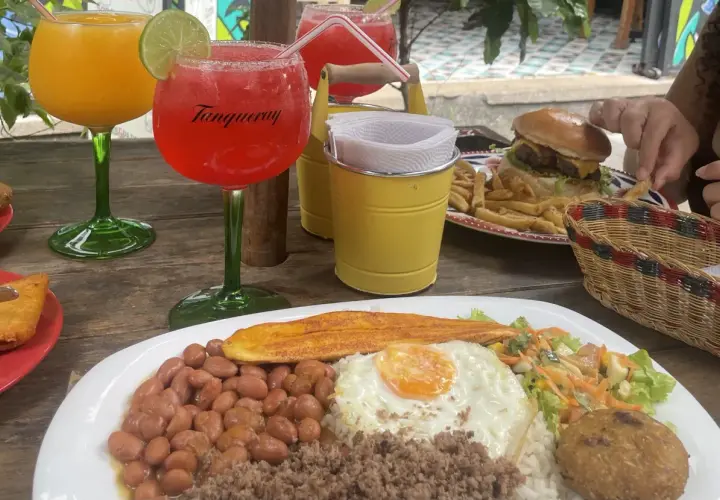 Bebidas y comida en una mesa de madera. Hay dos copas con bebidas, una hamburguesa con papas fritas, y un plato con arroz, frijoles, huevo frito y plátano. Es hora de pedir la cuenta en inglés.