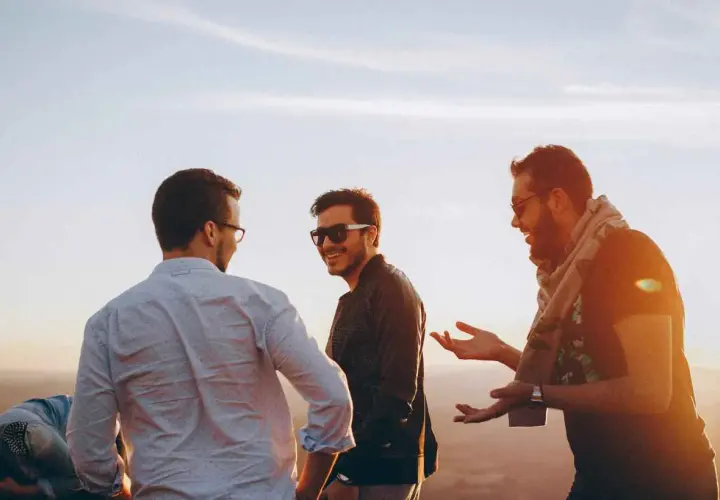 Tres amigos riendo y conversando al atardecer. Uno dice: "Encantado de conocerte en inglés", mientras los otros dos escuchan atentamente.