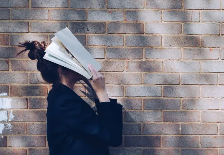 Persona con un libro abierto cubriendo su rostro, apoyada contra una pared de ladrillos. Parece estar inmersa en la lectura, quizás buscando un momento de tranquilidad.