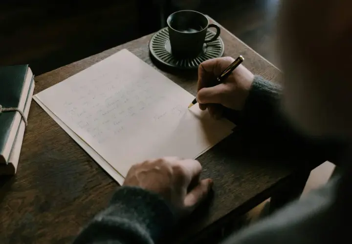 Persona escribe una carta en inglés, con una taza de café y un libro al lado.