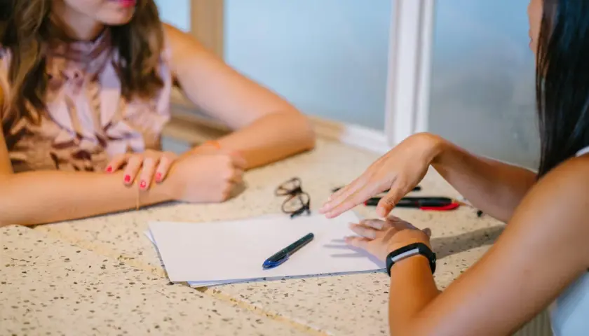 Dos mujeres discuten un proyecto sobre una mesa con papeles y bolígrafos. "Yo también," dice una, señalando.