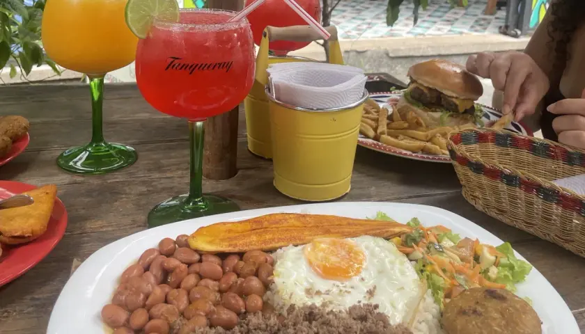 Bebidas y comida en una mesa de madera. Hay dos copas con bebidas, una hamburguesa con papas fritas, y un plato con arroz, frijoles, huevo frito y plátano. Es hora de pedir la cuenta en inglés.