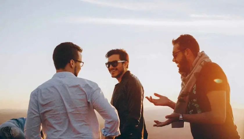 Tres amigos riendo y conversando al atardecer. Uno dice: "Encantado de conocerte en inglés", mientras los otros dos escuchan atentamente.