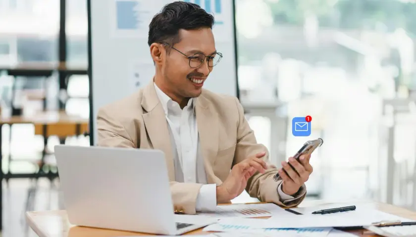 Hombre sonríe revisando su móvil con notificación de email. Busca ejemplos de email formal en inglés para su trabajo.