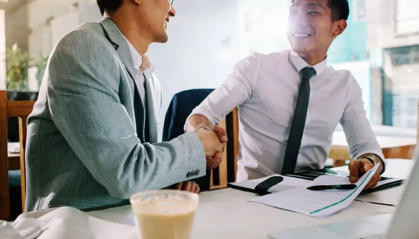 Dos hombres de negocios asiáticos se dan la mano en una cafetería. Uno sonríe, el otro asiente. Gracias por avisar en inglés.