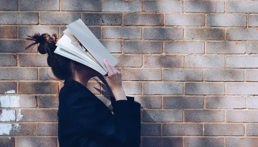Persona con un libro abierto cubriendo su rostro, apoyada contra una pared de ladrillos. Parece estar inmersa en la lectura, quizás buscando un momento de tranquilidad.