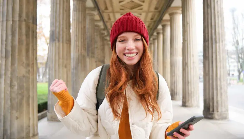 Joven sonríe con un teléfono en la mano, lista para enviar su carta de motivación Erasmus.