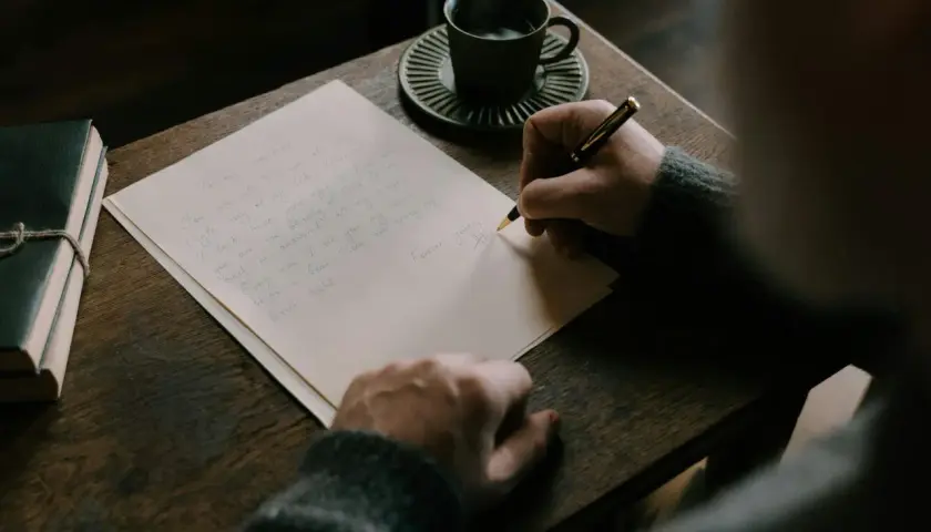 Persona escribe una carta en inglés, con una taza de café y un libro al lado.