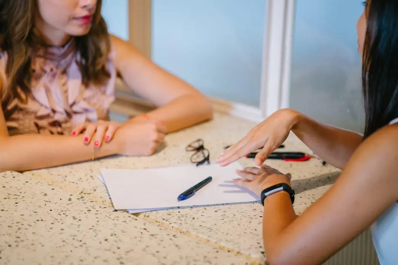 Dos mujeres discuten un proyecto sobre una mesa con papeles y bolígrafos. "Yo también," dice una, señalando.