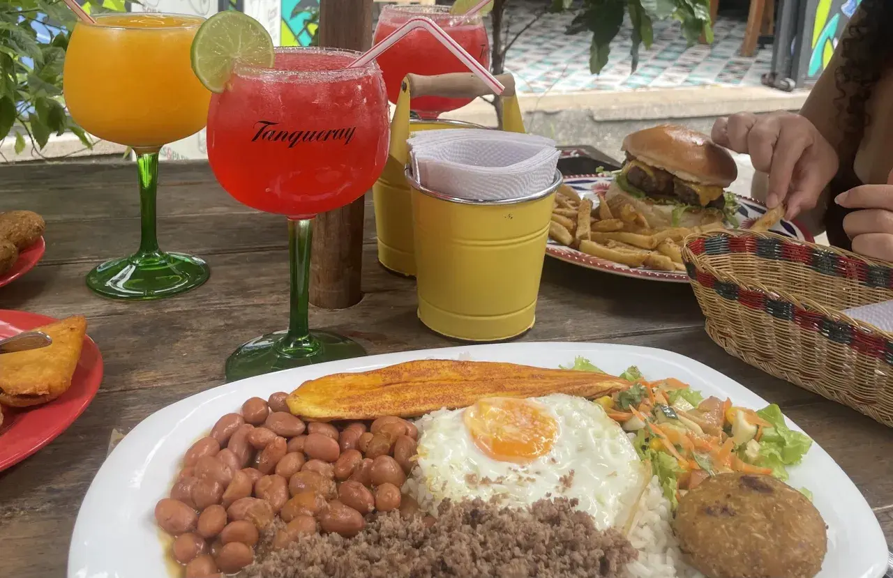 Bebidas y comida en una mesa de madera. Hay dos copas con bebidas, una hamburguesa con papas fritas, y un plato con arroz, frijoles, huevo frito y plátano. Es hora de pedir la cuenta en inglés.