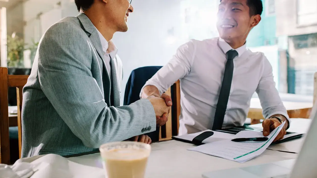 Dos hombres de negocios asiáticos se dan la mano en una cafetería. Uno sonríe, el otro asiente. Gracias por avisar en inglés.