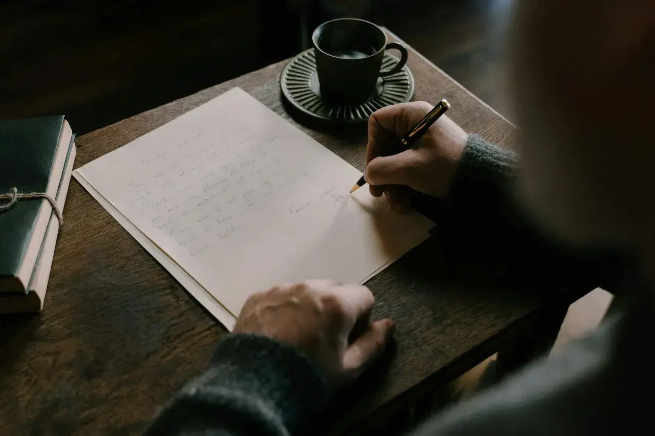Persona escribe una carta en inglés, con una taza de café y un libro al lado.