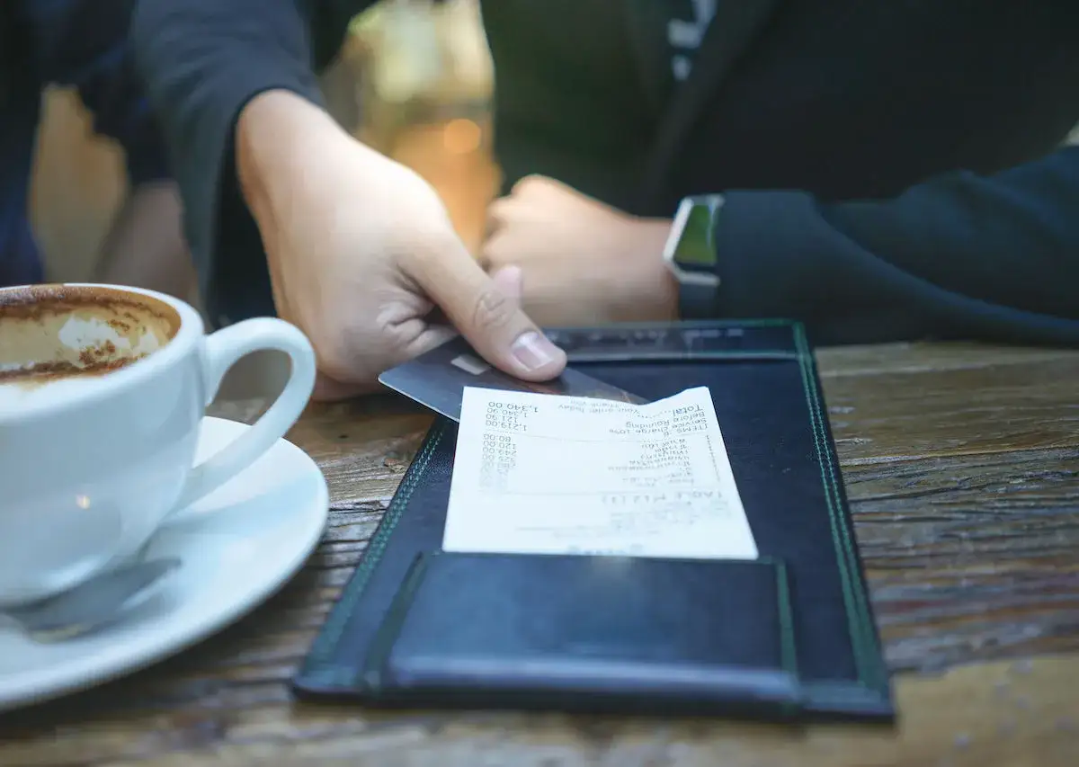 Mano con tarjeta de cr&eacute;dito lista para pagar la cuenta en ingl&eacute;s, junto a una taza de caf&eacute;.
