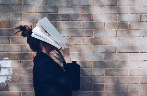 Persona con un libro abierto cubriendo su rostro, apoyada contra una pared de ladrillos. Parece estar inmersa en la lectura, quizás buscando un momento de tranquilidad.